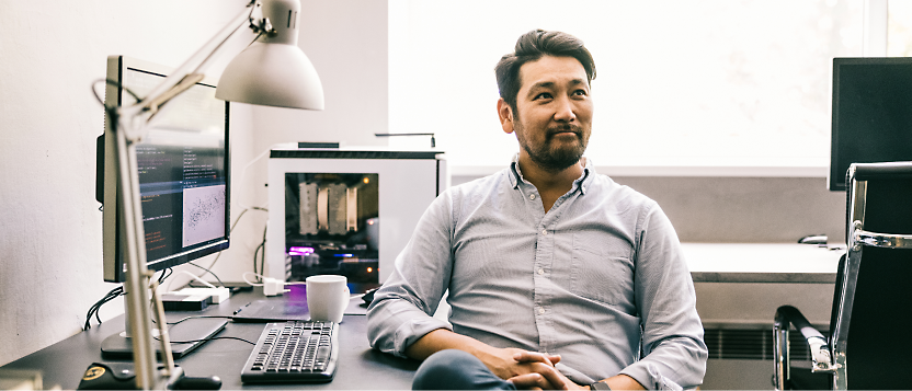 Hombre asiático con una camiseta azul sentado en un escritorio con equipos y mirando cuidadosamente a un lado en una oficina bien iluminada.
