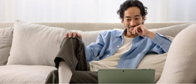 En person som sitter på en sofa og ser på en bærbar datamaskin med et lite smil, iført en lyseblå skjorte og mørke bukse.