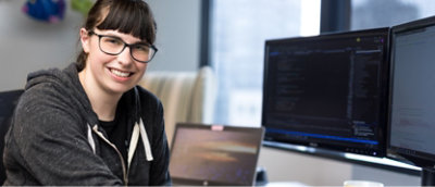 En person som bruker briller og en hettegenser smiler mens han sitter ved et skrivebord med flere dataskjermer som viser kode.