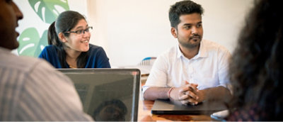 To personer sitter ved et bord og diskuterer noe. Den ene smiler og bruker briller.