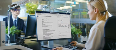 To personer i et kontormiljø: person i forgrunnen som arbeider på en stasjonær datamaskin som viser et detaljert grensesnitt.