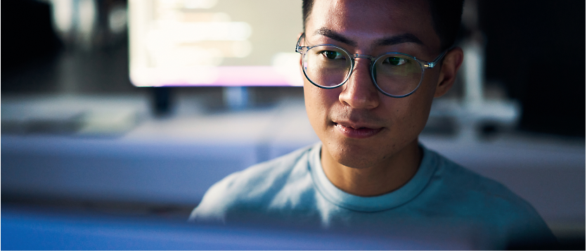 Loş bir odada odaklanmış bir şekilde bilgisayarda çalışan ve yüzüne odaklanılmış gözlüklü, genç Asyalı adam