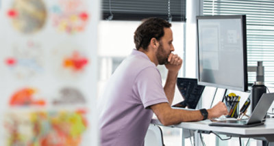 Fotografie osoby sedící u stolu v atriu pracující na přenosném počítači Na pozadí jsou lidé, kteří se procházejí.