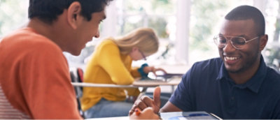 Two people sitting at a table, smiling and interacting, with another person in the background focused on a tablet.