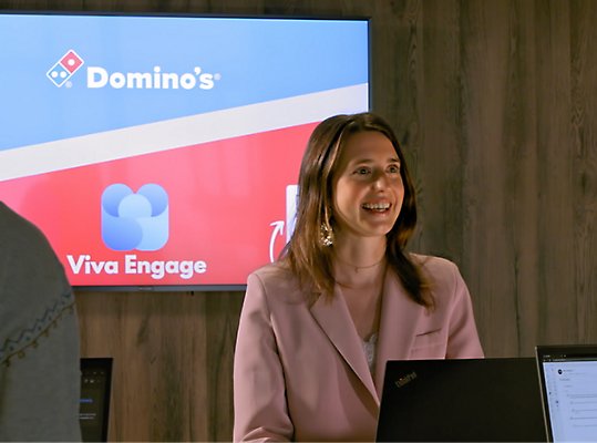 Three people conversing at a counter under a screen displaying logos for Domino's and Viva Engage.