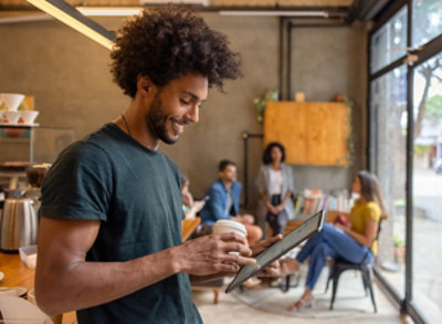 A team member looking at a tablet with a drink in hand, team members casually collaborating in the background.