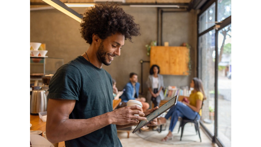 A team member looking at a tablet with a drink in hand, team members casually collaborating in the background.