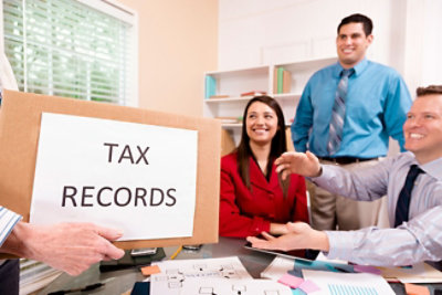 Četiri osobe u kancelariji, od kojih jedna drži kutiju sa natpisom „PORESKE EVIDENCIJE“, a ostale tri sede oko stola na kojem se nalaze papiri 