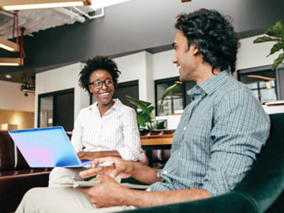 Two colleagues collaborate in a modern office lounge, using a laptop and mobile device.