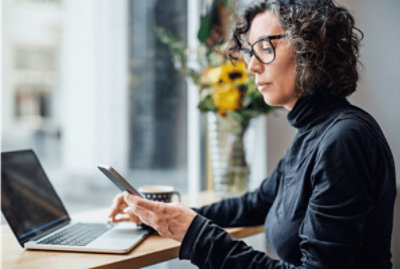 机に向かって座り、ノート PC を使用しながら携帯電話を見ている人。