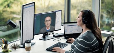 ヘッドフォンを付けて机に座り、複数の画面を使用して Teams のビデオ会議に参加している人。