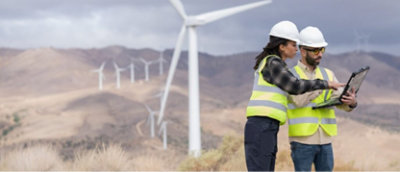 Two construction workers looking at a laptop in front of wind turbines.