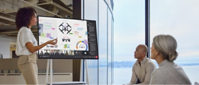 A person presenting drone-related content on a digital screen to two seated individuals in a modern office setting.