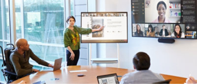 A woman presents at a conference table, pointing to a screen displaying slides.