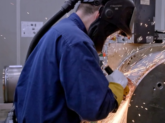 A welder wearing protective face gear and welding a large piece of machinery.