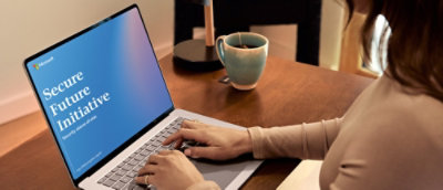  A person using a laptop displaying a 'Secure Future Initiative' presentation while working at a wooden desk with a coffee mug.
