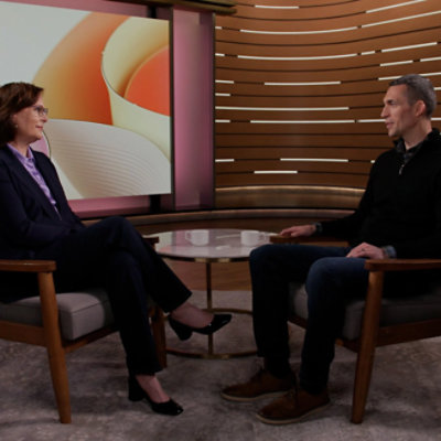 Two people sitting in armchairs facing each other, engaged in conversation. A modern, abstract backdrop is behind them.