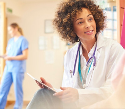 Un professionnel de la santé en blouse blanche avec un stéthoscope regardant une tablette, avec un autre membre du personnel médical en arrière-plan.