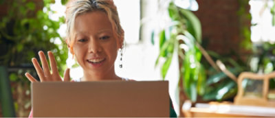 明るい部屋に座り、植物を背景にノート PCの画面に向かって微笑みながら手を振る人。