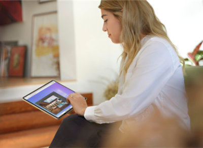 A person with long hair uses a stylus on a tablet, sitting on a sofa in a well-lit room with art pieces in the background.