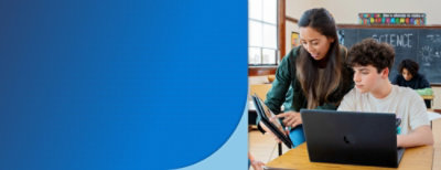 Eine Frau zeigt einem Schüler, der vor einem Laptop in einem Klassenzimmer sitzt, etwas auf einem Tablet.