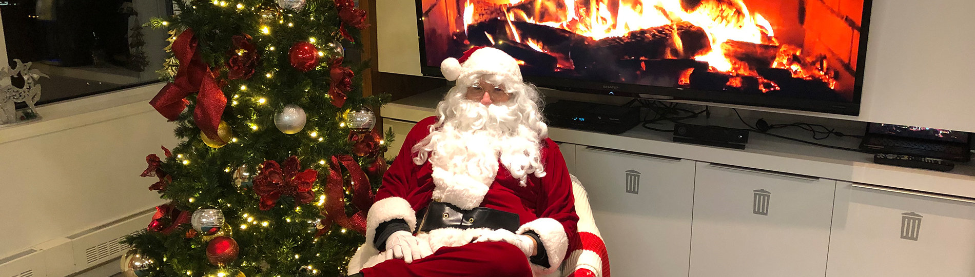 Gilles Garceau dressed as Santa Claus, sitting by digital fireplace and Christmas tree