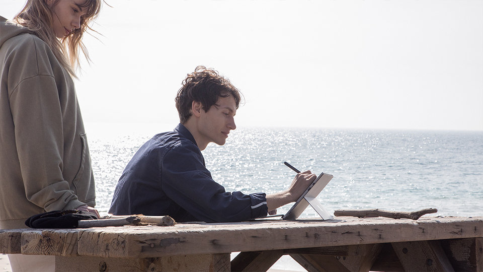 A man uses Surface Pro 5th Gen for Business on a bench near the ocean.