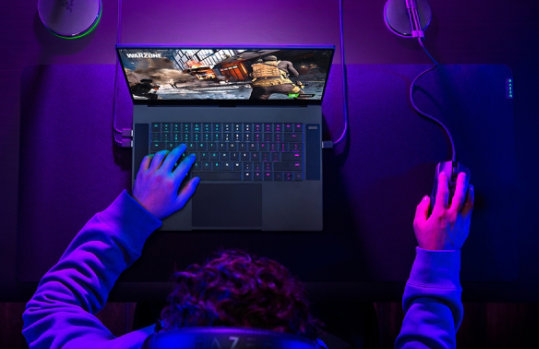 Man gaming on Razer Blade 15 Gaming Laptop with full desktop setup and gaming peripherals.