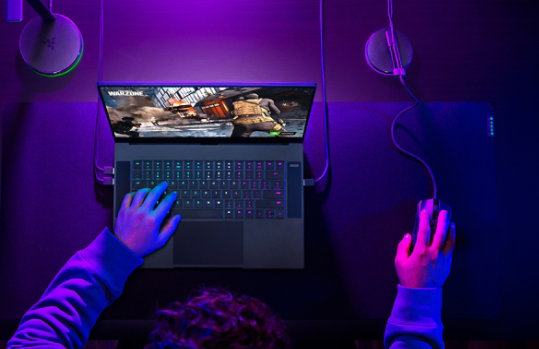 A person gaming on a Razer Blade 15 with full desktop setup and accessories including a mouse.