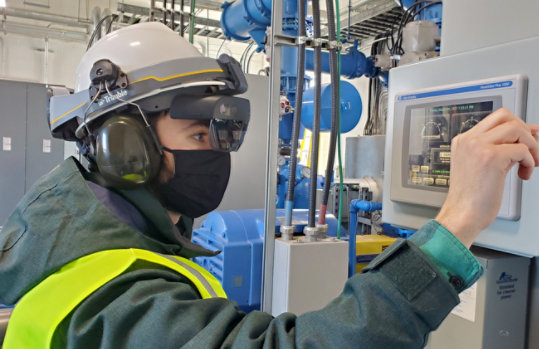 A person wears a face mask with their Trimble XR 10 with HoloLens 2 in an industrial environment while interacting with a screen.