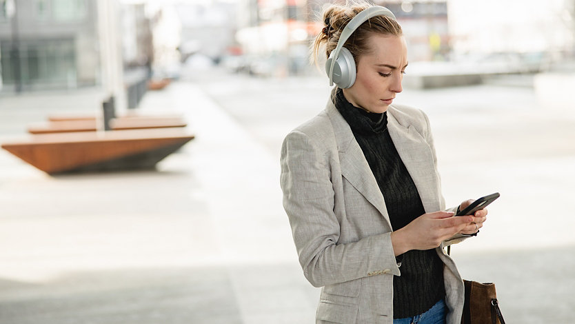 A woman using Bose Noise Cancelling Headphones 700 UC in Silver with her phone outdoors.