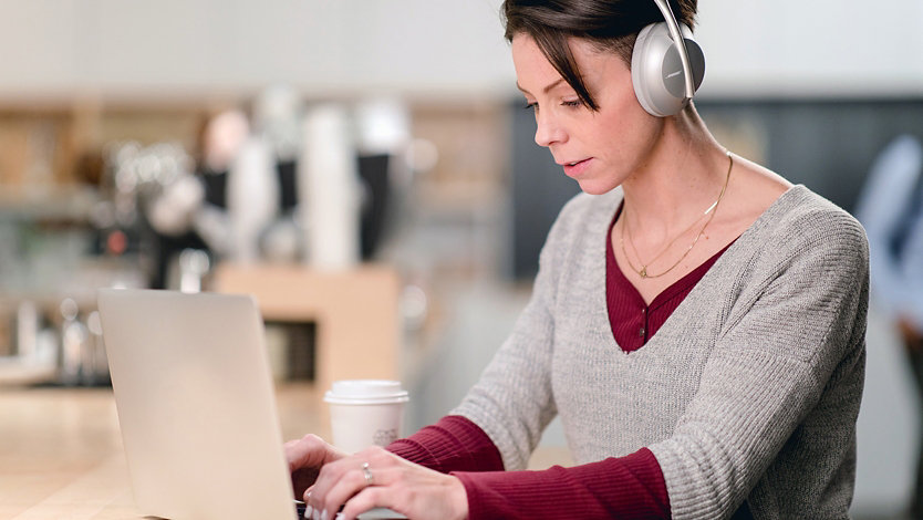 A woman using Bose Noise Cancelling Headphones 700 UC in Silver with her laptop in a café setting.