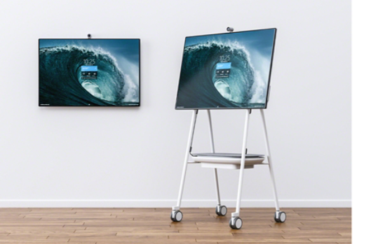 Ein Surface Hub 2S an der Wand neben einem Surface Hub 2S auf einem Steelcase Roam-Geräteständer