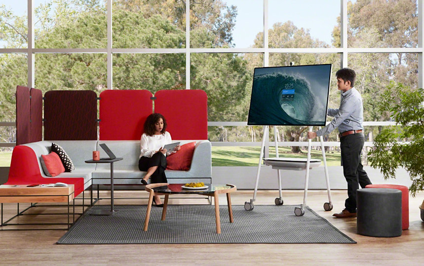 Un hombre de pie junto a un dispositivo Surface Hub 2S mientras una mujer sentada en un sofá le mira