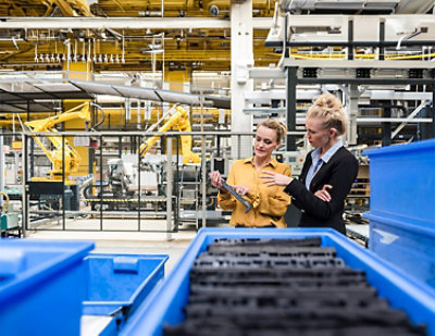 Two women are discussing a component in an industrial factory setting surrounded by machinery and equipment. 
