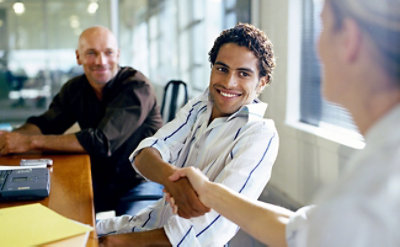 Dve osobe se rukuju za stolom tokom sastanka, a treća osoba posmatra i osmehuje se.