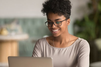 A person with short curly hair and glasses is smiling while using a laptop.