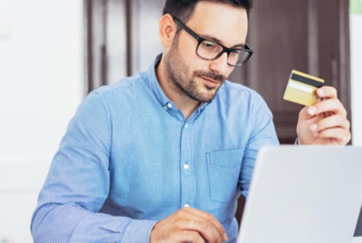 Ένας άντρας φοράει γυαλιά και κάθεται σε ένα γραφείο, κρατώντας μια πιστωτική κάρτα στο ένα χέρι και πληκτρολογώντας σε έναν φορητό υπολογιστή με τον άλλο.