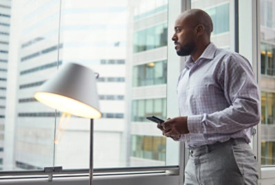Un homme vêtu d’une chemise à carreaux claire se tient dans un bureau, un smartphone à la main et regarde par la fenêtre 