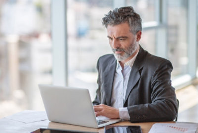 A man with gray hair and a beard sits at a wooden desk typing on a laptop in a bright office with large windows.