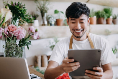 Una florista sonriente, con una camiseta blanca y un delantal, mira una tableta en una tienda llena de varias plantas y flores en macetas.