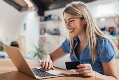 A smiling woman holding a credit card is using a laptop at a table in a bright room.