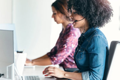 Three people at desks working on laptops and wearing headsets in a bright, modern office setting. 