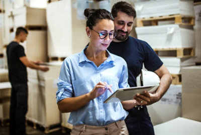 仓库中的两个人正在查看平板电脑屏幕。背景是一个人站纸堆旁边。