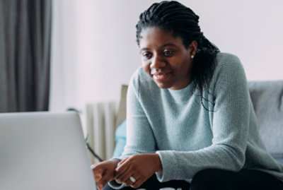 A person with braided hair sits on a couch and looks at a laptop screen in a well-lit room.