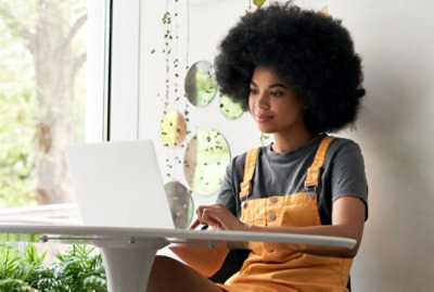 Una persona con cabello rizado sentada en una mesa redonda trabajando en un portátil; viste una camisa gris y un mono amarillo