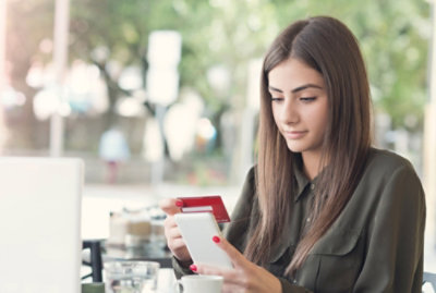 A woman sits at an outdoor table using a smartphone and holding a credit card, with a laptop and coffee cup in front of her.