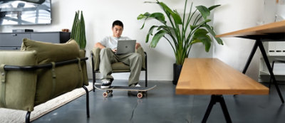 Osoba siedzi na krześle w nowoczesnym salonie, korzystając z laptopa umieszczonego na kolanach. Pod stopami leży deskorolka, a w tle widać rośliny.