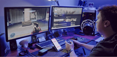 A person sitting their desk playing a first person shooter video game on a computer monitor.