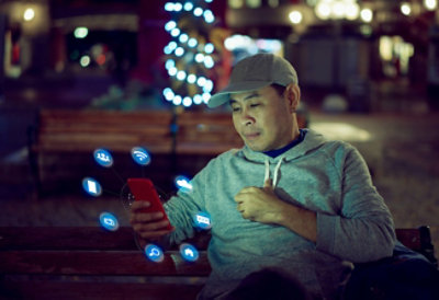 Muškarac u sivoj dukserici sa kapuljačom i sa kapom na glavi sedi noću na klupi i koristi pametni telefon sa hologramskim ikonama koje predstavljaju različite aplikacije